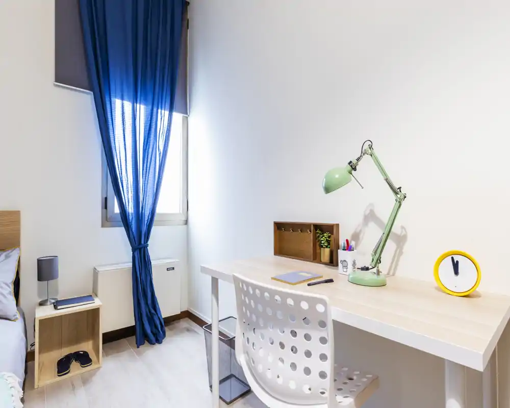 Bright, modern bedroom corner with a single bed visible at left and a tidy study desk with lamp and chair on the right; natural light through a window with blue curtains.