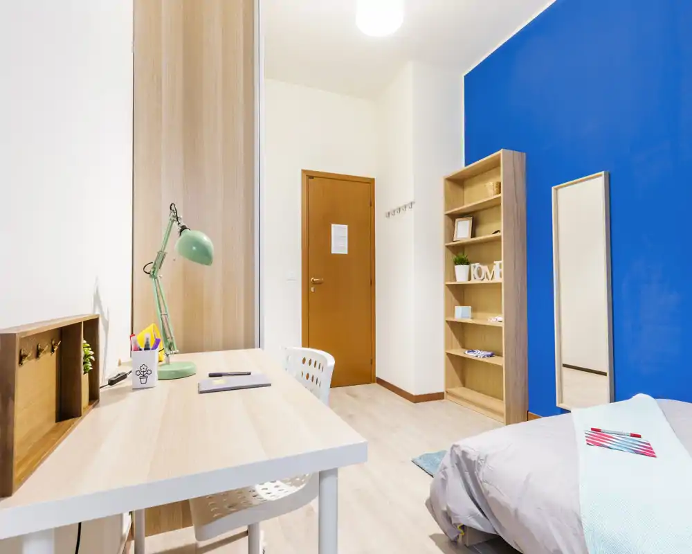 Bright, modern bedroom with a study desk, bookshelf and a bold blue accent wall. The neatly made bed and clean wood finishes create a welcoming, functional space ideal for renters or students.