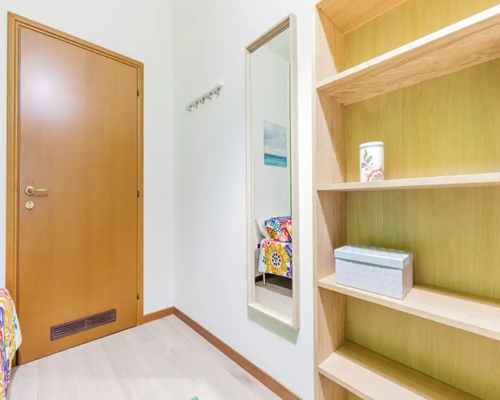 Angolo di camera da letto con porta in legno, specchio a muro e scaffalatura integrata; il colorato copriletto è visibile nello specchio, indicando un letto singolo nelle vicinanze. Luminoso, ordinato e arredato in modo semplice, ideale per un affitto compatto.
