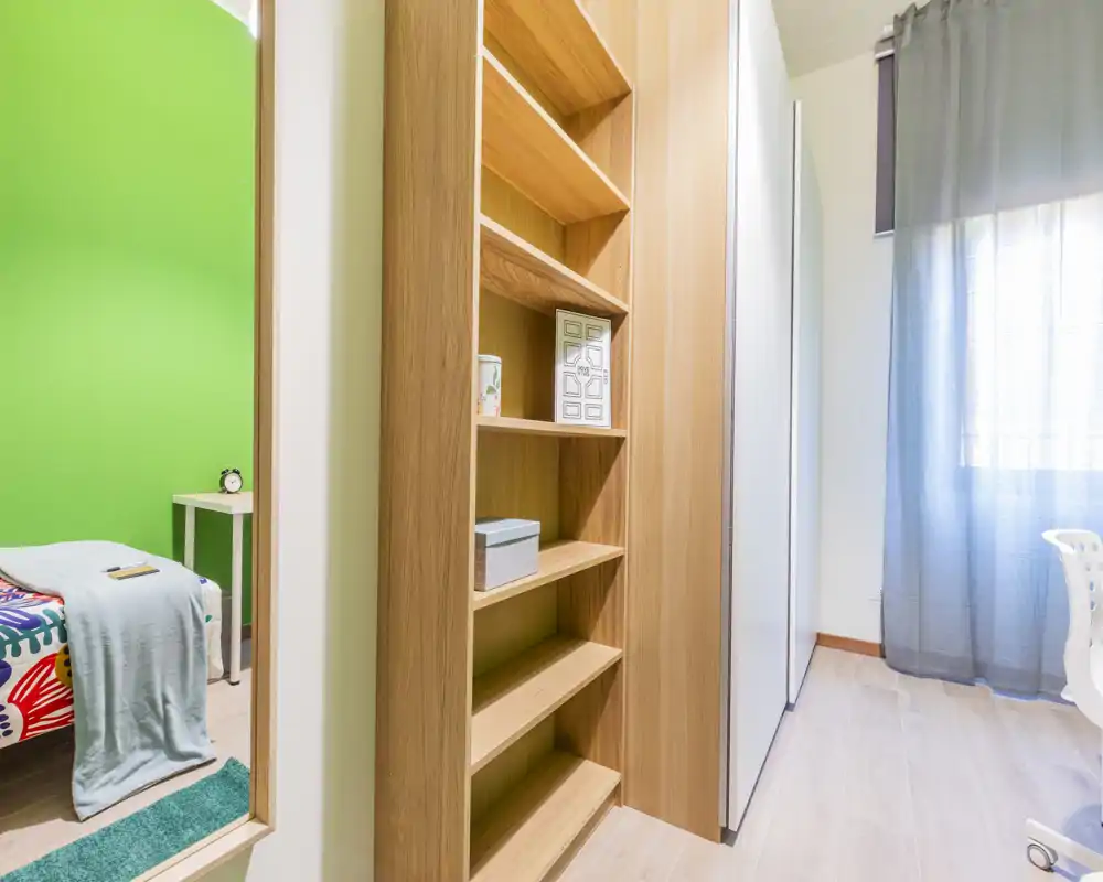 Angolo di camera da letto luminoso e moderno con un letto singolo colorato visibile attraverso la porta, una libreria in legno e un armadio vicino a una finestra con tende leggere.