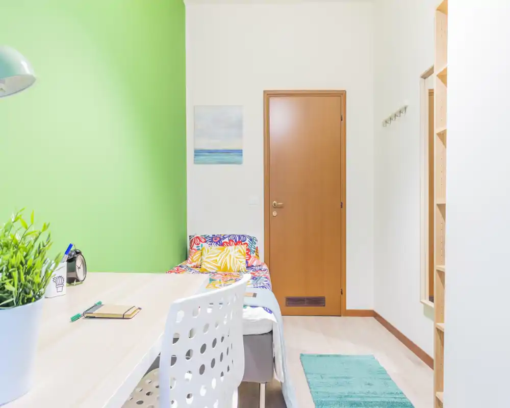 Camera singola compatta e luminosa con letto colorato, angolo scrivania e pavimento in legno chiaro — ambiente fresco, ben illuminato e curato, ideale per affitti brevi.