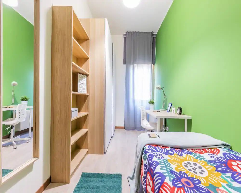 Camera da letto singola luminosa con copriletto colorato, scaffalatura integrata e una piccola scrivania vicino alla finestra — ideale per uno studente o un occupante singolo.