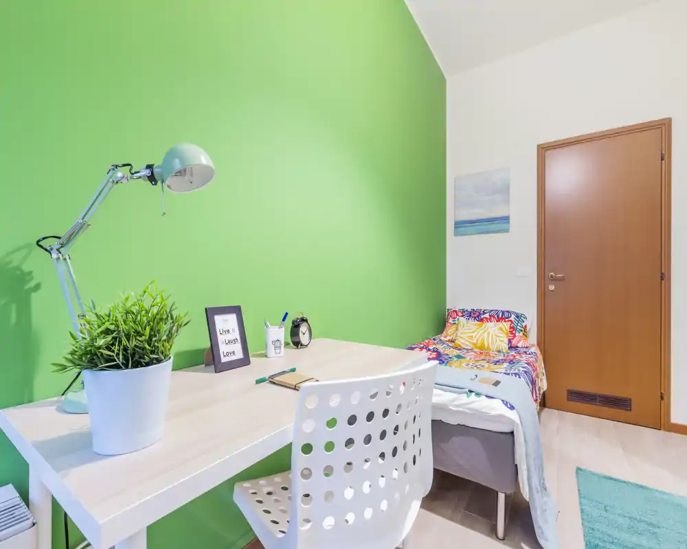 Camera da letto compatta e luminosa con angolo studio. Il letto singolo con biancheria colorata è accanto a una scrivania bianca ordinata su una parete verde brillante — ideale per studenti o affitti brevi.
