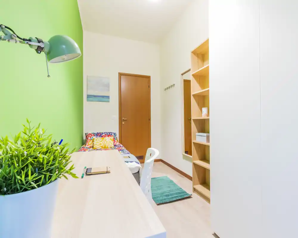 Camera singola compatta e luminosa con scrivania lungo una parete verde, biancheria colorata e scaffalatura integrata — ideale per studenti o affitti brevi.