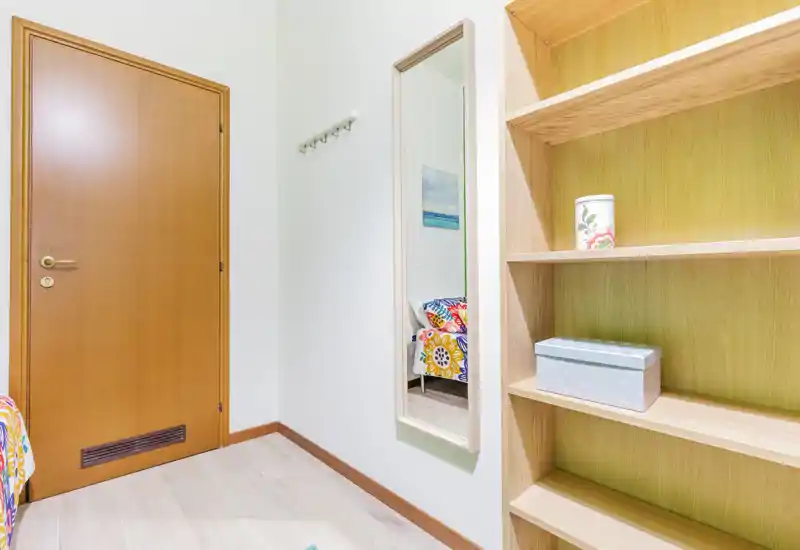 Rincón de dormitorio con puerta de madera, espejo de pared y estanterías empotradas; la ropa de cama colorida se ve en el espejo indicando una cama individual cercana. Luminoso, ordenado y con mobiliario sencillo, ideal para un alquiler compacto.