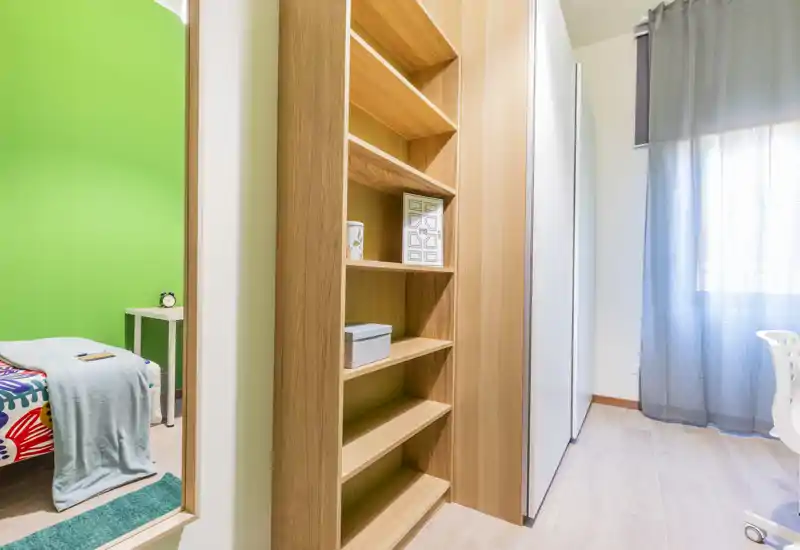Rincón de dormitorio luminoso y moderno con una cama individual colorida visible a través de la puerta, una estantería de madera y un armario junto a una ventana con cortinas translúcidas.