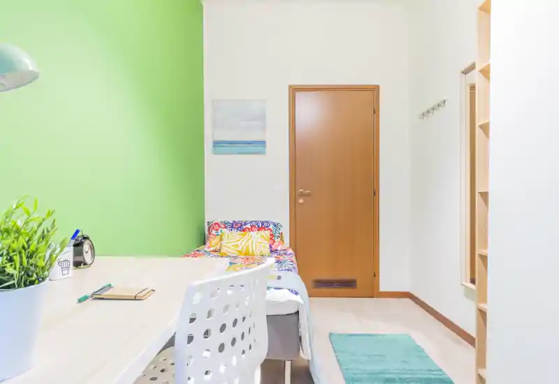Habitación individual compacta y luminosa con cama colorida, zona de escritorio y suelo de madera clara — ambiente fresco, bien iluminado y cuidado, ideal para alquileres de corta estancia.