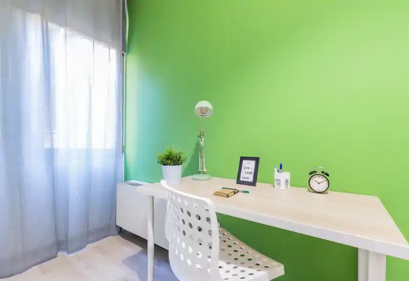 Rincón de oficina en casa luminoso y compacto con escritorio de madera clara, silla blanca moderna, planta decorativa y lámpara de lectura frente a una pared verde intensa—ideal para trabajar con concentración.