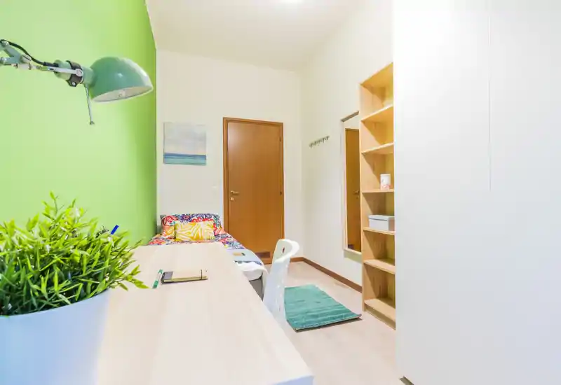 Habitación individual compacta y luminosa con escritorio junto a una pared verde, ropa de cama colorida y estanterías empotradas — ideal para estudiantes o alquileres de corta estancia.