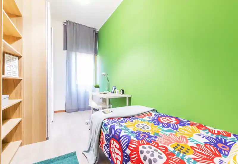Dormitorio individual luminoso con colcha floral colorida, pared verde, escritorio y estanterías — ideal para un estudiante o una persona sola.