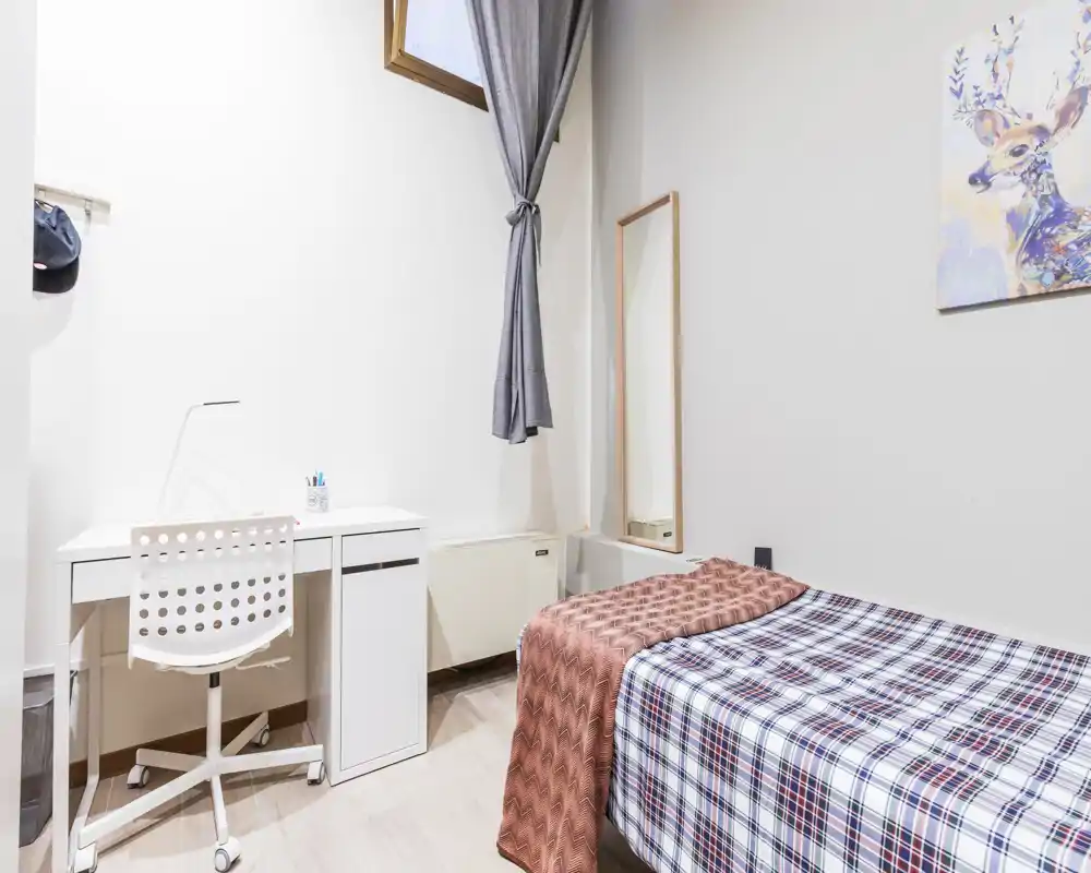 Camera luminosa e compatta con letto singolo, scrivania ordinata e specchio alto — ideale per studenti o soggiorni brevi. Arredo minimal, luce naturale e pavimento in legno chiaro.