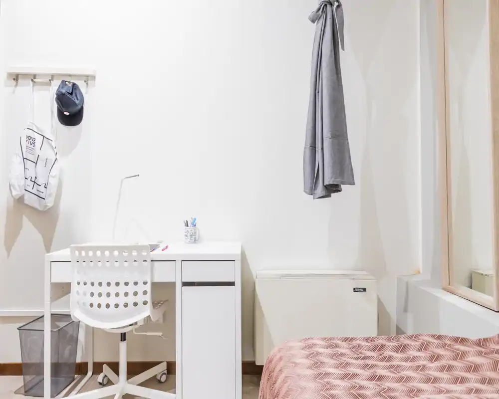 Angolo camera luminoso e minimale con scrivania bianca, sedia e letto parzialmente visibile con copriletto a motivo. Spazio pulito e ben illuminato ideale per soggiorni concentrati.