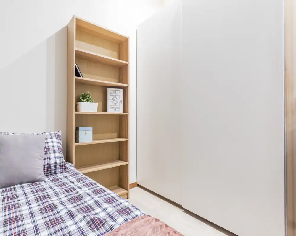 Angolo di camera singola luminoso e moderno con copriletto a quadri, libreria in legno alta e grande armadio bianco — ambiente pulito e accogliente ideale per un singolo.