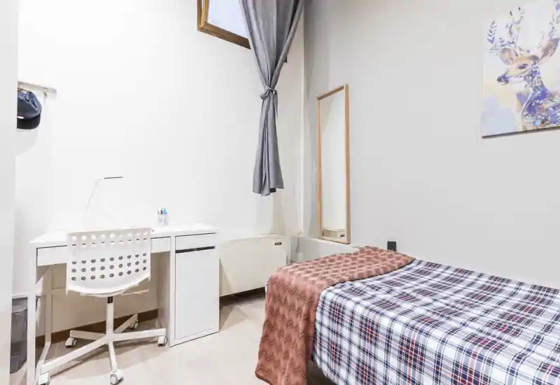 Camera luminosa e compatta con letto singolo, scrivania ordinata e specchio alto — ideale per studenti o soggiorni brevi. Arredo minimal, luce naturale e pavimento in legno chiaro.