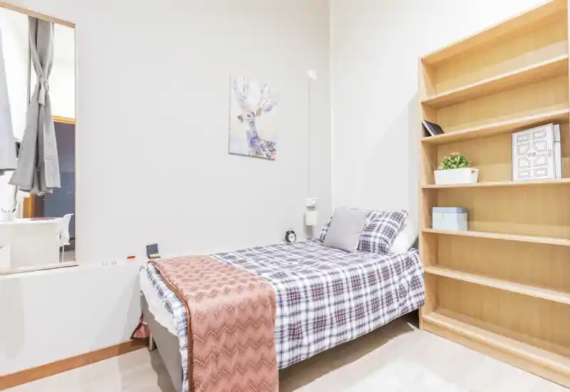 Camera da letto singola luminosa e ben allestita con copriletto a quadri, plaid accogliente e mensole aperte — perfetta per un annuncio che valorizza uno spazio notte compatto e illuminato.