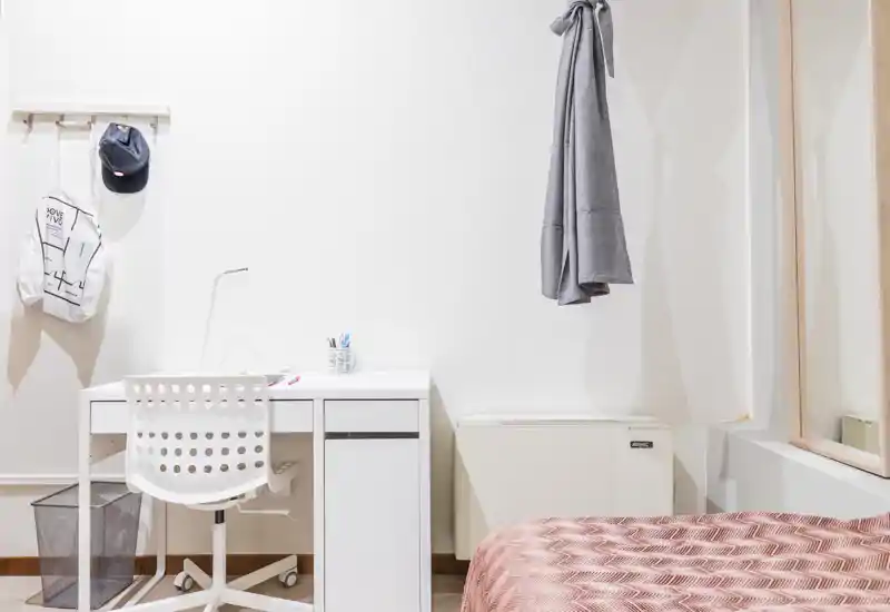 Rincón de dormitorio luminoso y minimalista con escritorio blanco, silla y cama parcialmente visible con edredón estampado. Paredes blancas limpias, área de descanso funcional y zona de trabajo práctica para alquileres.