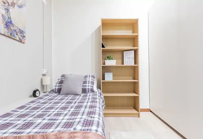 Camera singola luminosa e ordinata con copriletto a quadretti, cuscino morbido e alta libreria in legno. Lo spazio è pulito e accogliente, con luce piacevole e arredamento essenziale.