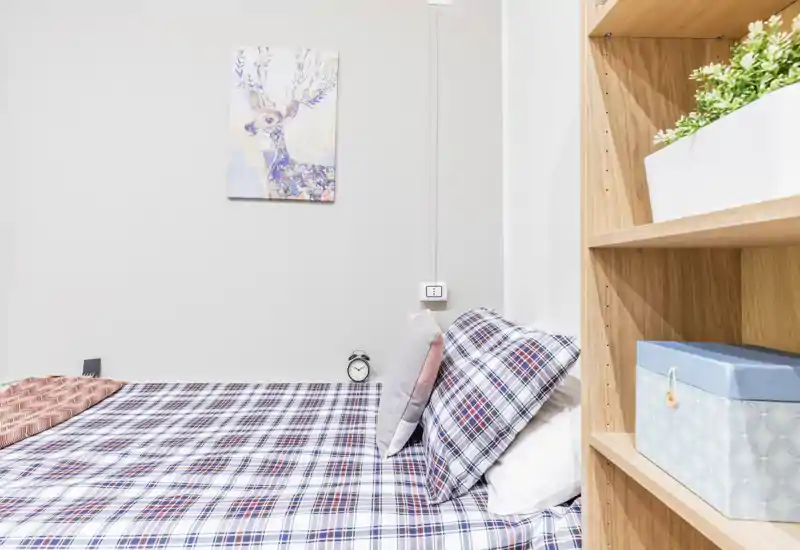 Camera da letto luminosa e ordinata con letto coperto da plaid a quadri, cuscini decorativi e una libreria in legno a destra; arredo minimale e un quadro a parete che rendono l'ambiente accogliente.