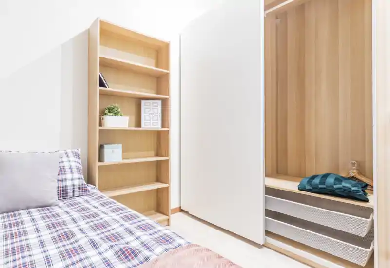 Dormitorio luminoso y minimalista con colcha a cuadros, almohada y armario abierto de madera; estanterías claras que aportan almacenamiento y sensación de amplitud.