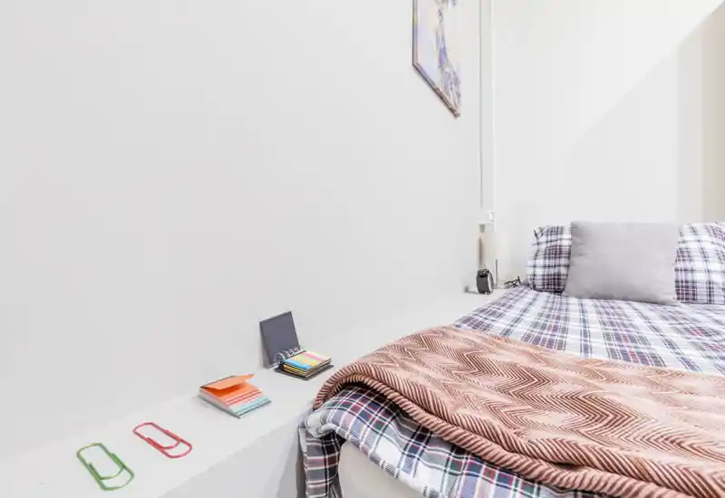 Rincón de dormitorio luminoso y ordenado con colcha a cuadros, almohada suave y manta plegada; paredes blancas y pequeños objetos decorativos crean un espacio para dormir moderno y sereno.