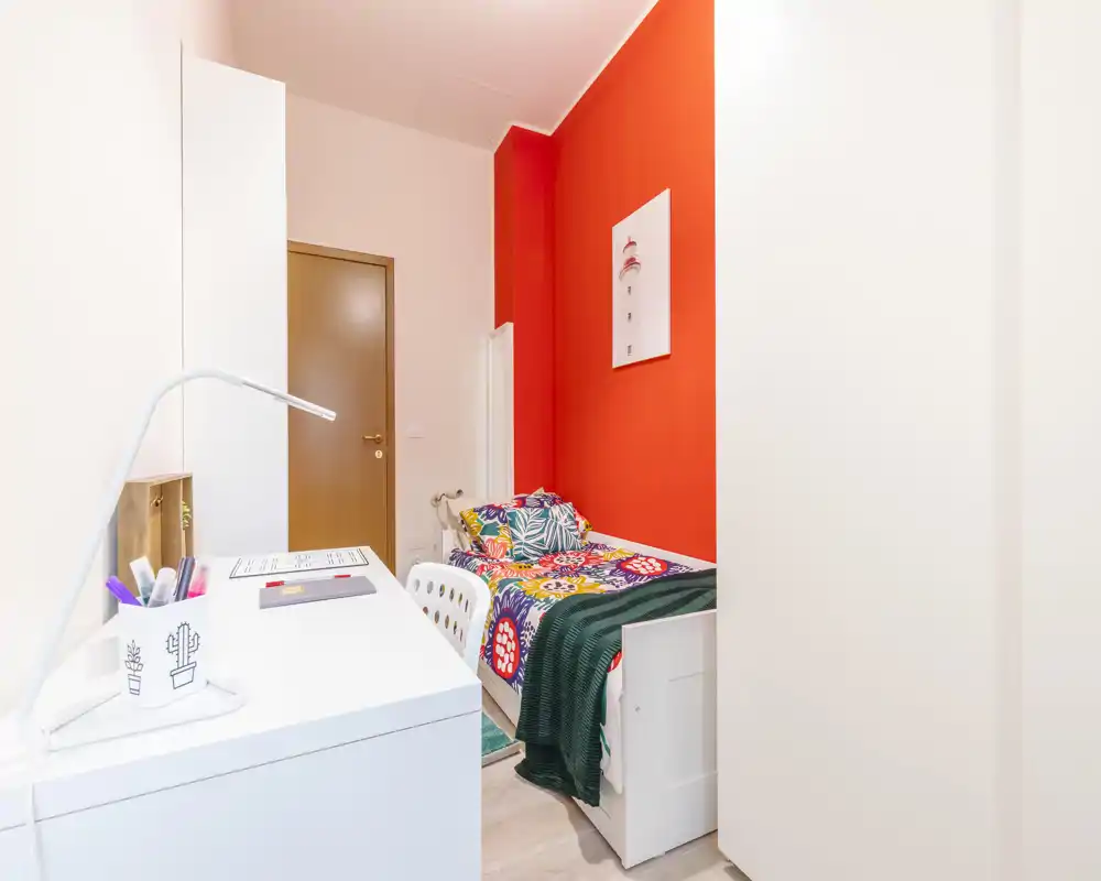Chambre compacte et colorée avec un lit simple, coussins décoratifs et un bureau bien rangé près du lit. Style moderne et lumineux avec un mur d'accent rouge et des rangements pratiques.