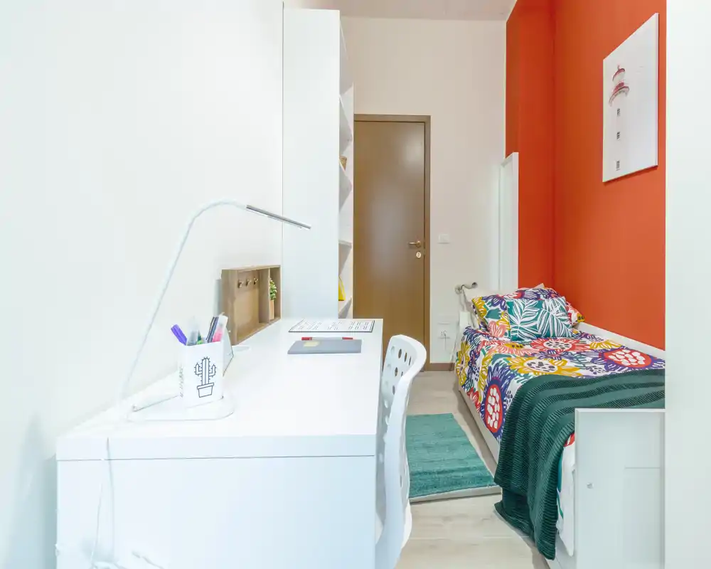 Chambre simple compacte et lumineuse avec une couette colorée, un bureau avec lampe et des étagères organisées — idéale pour un étudiant ou une personne seule.