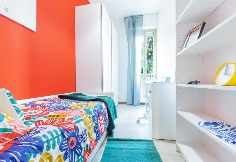 Chambre simple lumineuse et colorée avec une couette à motifs, tapis turquoise et belle lumière naturelle depuis la fenêtre; étagères rangées et petit bureau, parfaite pour un étudiant ou un locataire seul.