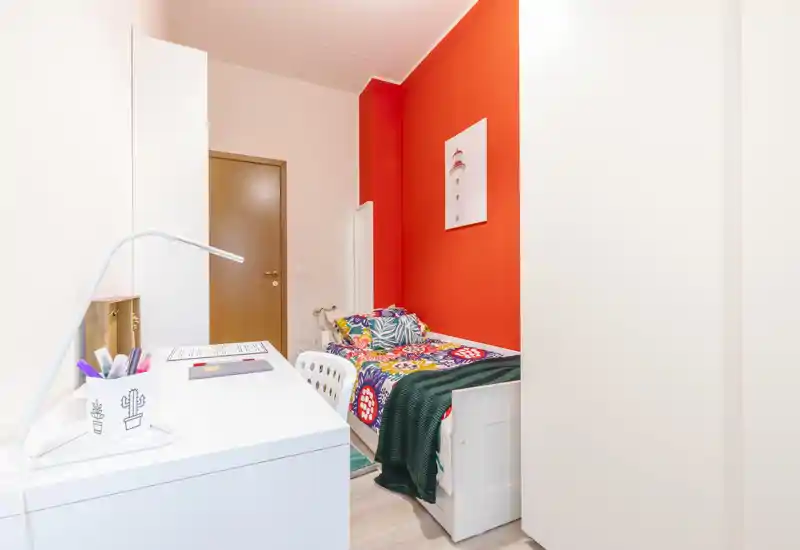Chambre compacte et colorée avec un lit simple, coussins décoratifs et un bureau bien rangé près du lit. Style moderne et lumineux avec un mur d'accent rouge et des rangements pratiques.