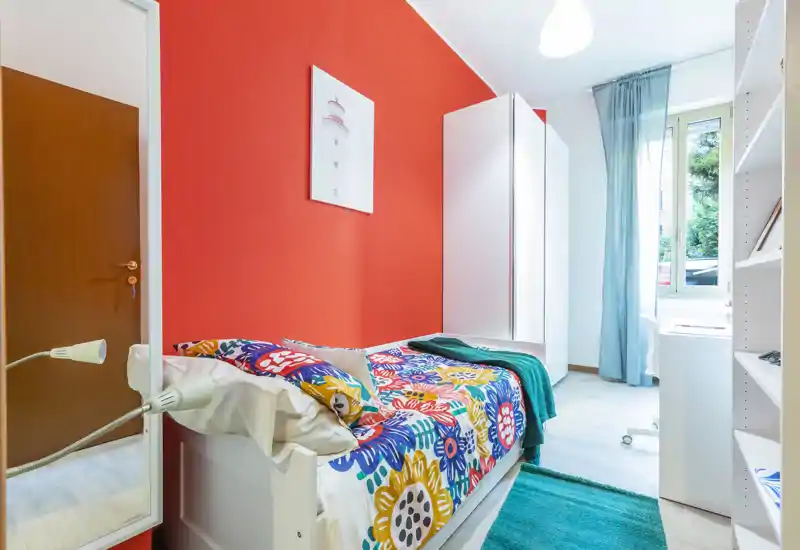 Chambre simple lumineuse et colorée avec couette à motifs, armoire blanche, bureau près de la fenêtre et textiles confortables. La lumière naturelle et la décoration vive rendent la pièce chaleureuse et accueillante.