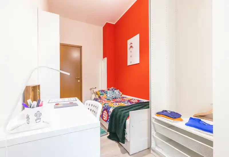 Chambre simple lumineuse et colorée avec bureau et rangement intégré. Le lit présente une literie vive sur un mur rouge accentué, avec un espace de travail au premier plan — parfait pour un étudiant ou une personne seule.