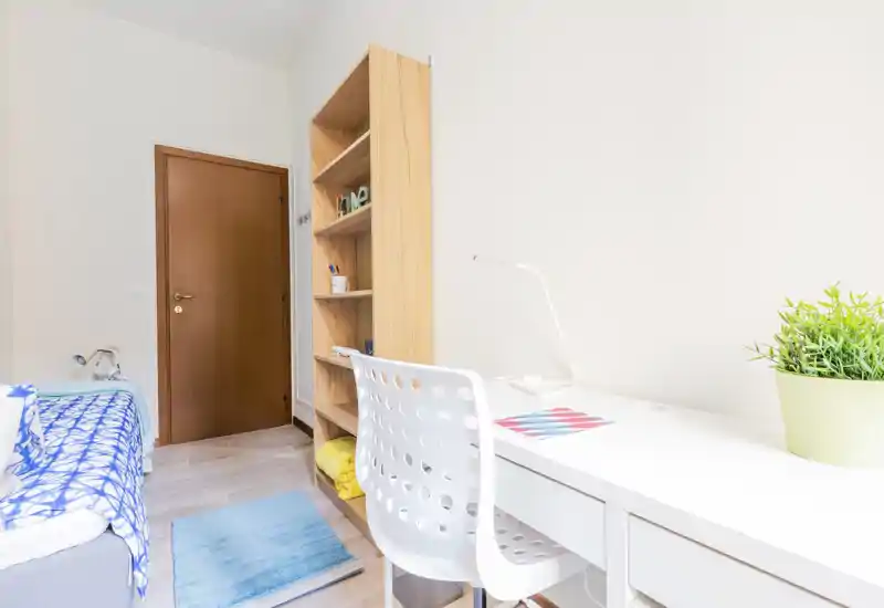 Dormitorio luminoso y ordenado con cama individual, estantería de madera y un escritorio blanco junto a la pared — ideal para estudiante o alquiler temporal.