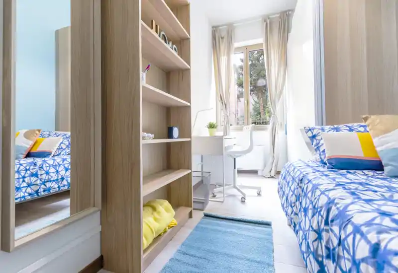 Camera singola luminosa e curata, con biancheria a fantasia, una scrivania vicino alla finestra e scaffalature integrate — perfetta per studenti o soggiorni brevi.