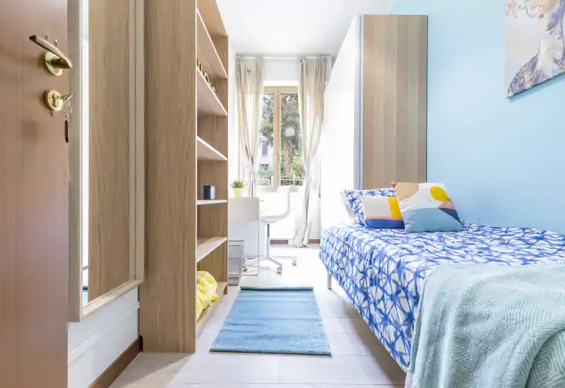 Camera da letto singola luminosa e ordinata con copriletto a fantasia, cuscini decorativi e una scrivania vicino alla finestra che illumina la stanza con luce naturale.