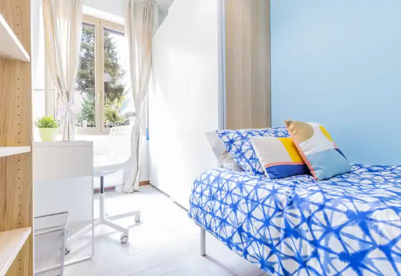 Camera da letto luminosa e compatta con letto singolo con biancheria blu a motivo, cuscini decorativi e una scrivania vicino alla finestra che lascia entrare molta luce naturale.