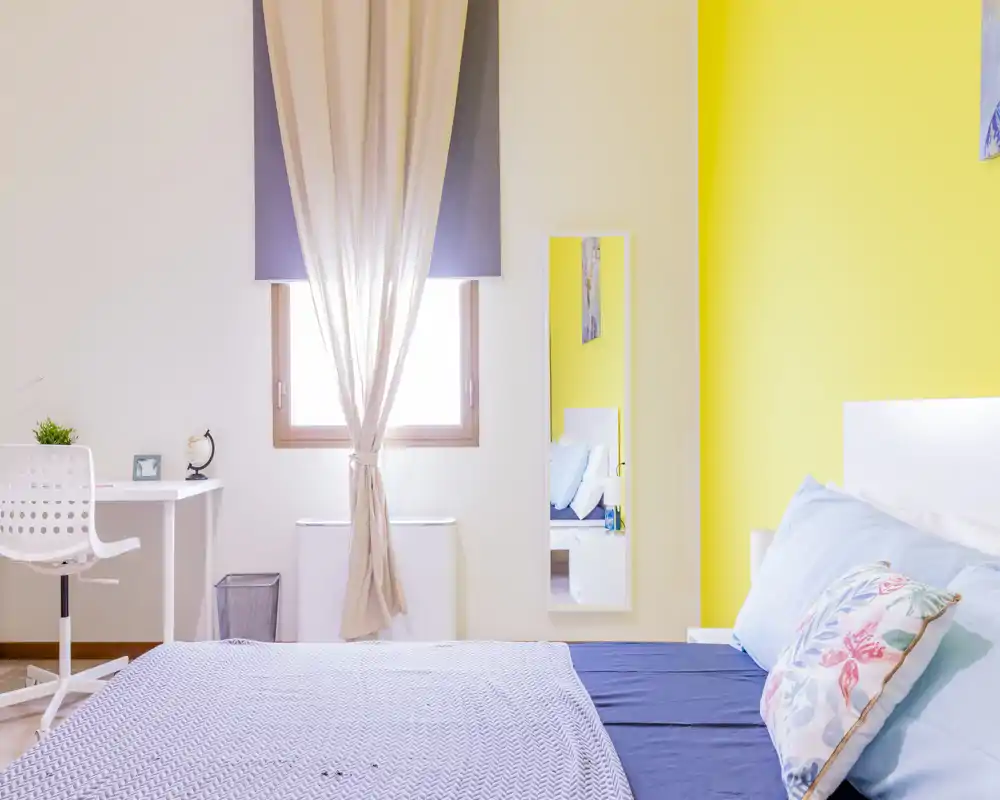 Bright, modern bedroom with a comfortable double bed, decorative pillows and a cheerful yellow accent wall; natural light from the window and a small desk make it ideal for short-term rentals.