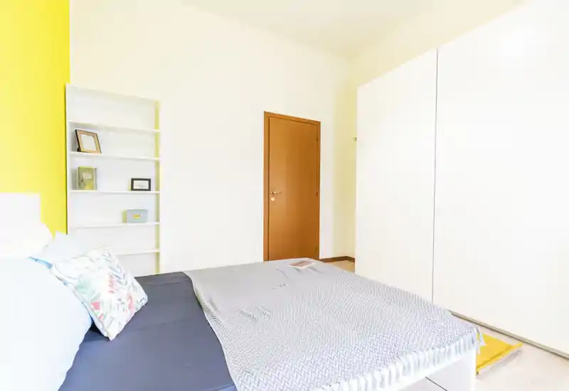 Camera da letto luminosa e moderna con un letto ampio, cuscini decorativi e un copriletto morbido; armadio bianco e parete gialla accentano l’ambiente pulito e arioso.