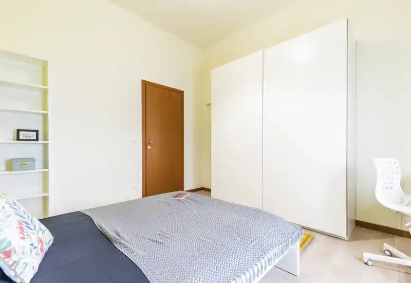 Camera da letto luminosa e minimal con grande armadio bianco, letto con cuscino decorativo e copriletto, scaffalatura e una sedia da scrivania — perfetta per un affitto ordinato.