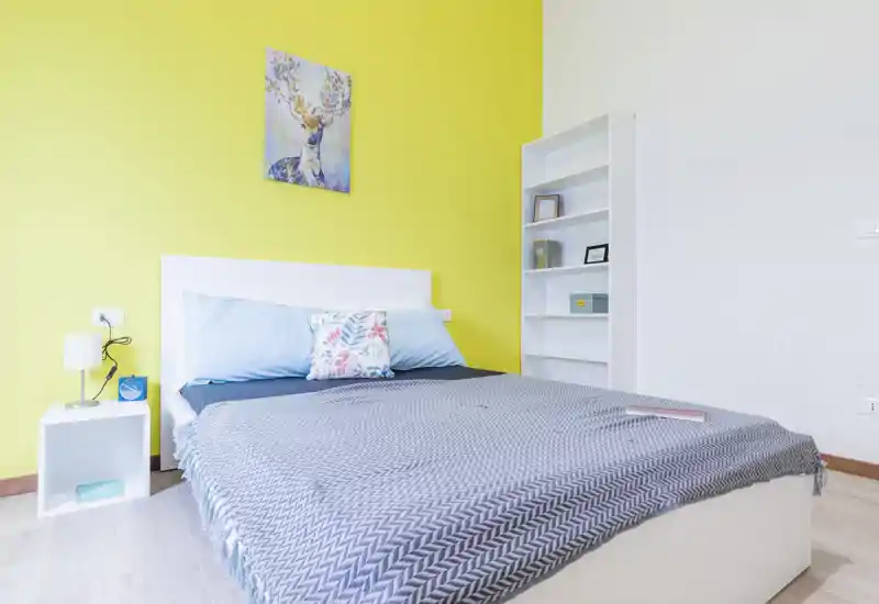 Camera da letto luminosa e moderna con letto matrimoniale davanti a una parete gialla, biancheria ordinata e una libreria bianca per riporre oggetti.