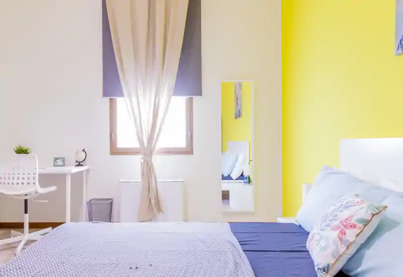 Camera da letto luminosa e moderna con letto matrimoniale, cuscini decorativi e una parete gialla vivace; luce naturale dalla finestra e una scrivania rendono lo spazio adatto ad affitti brevi.