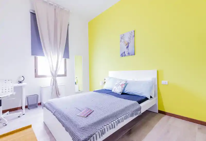 Camera da letto luminosa e moderna con letto bianco su piattaforma, cuscini decorativi e parete d'accento gialla. La luce naturale dalla finestra e i tessili morbidi creano un'atmosfera accogliente.