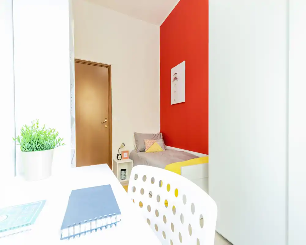 Camera da letto luminosa e compatta con letto singolo contro una parete rossa, piccolo comodino e una scrivania bianca in primo piano — ideale per studenti o soggiorni brevi.