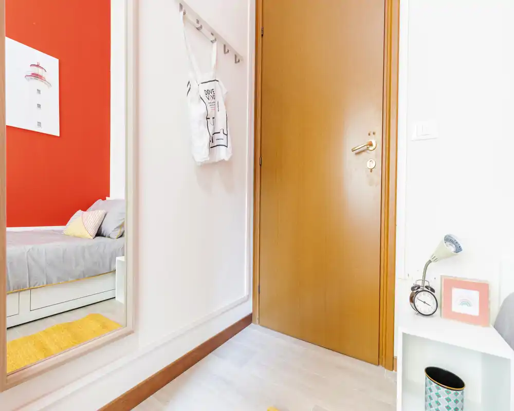 Ingresso luminoso con vista su una piccola camera: il letto è visibile attraverso la porta aperta e anche nello specchio a parete. Biancheria neutra, cuscini colorati e la porta in legno donano un ambiente semplice e accogliente.