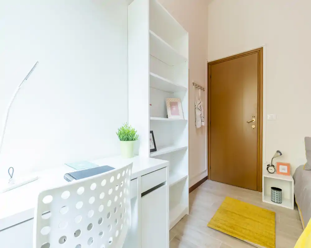Angolo camera luminoso e moderno con scrivania bianca, libreria e vista parziale del letto. Pavimento chiaro in legno, piccolo tappeto giallo e comodino per una soluzione comoda per una persona.