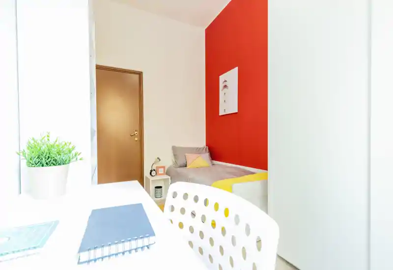 Camera da letto luminosa e compatta con letto singolo contro una parete rossa, piccolo comodino e una scrivania bianca in primo piano — ideale per studenti o soggiorni brevi.