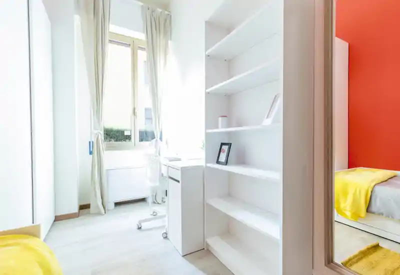 Camera singola luminosa e moderna con scrivania vicino alla finestra, scaffalatura bianca e tocchi gialli caldi. La luce naturale illumina la stanza e il letto ben ordinato visibile a destra rende l'ambiente accogliente.