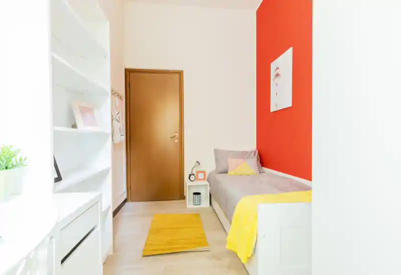 Camera singola luminosa e ben arredata con struttura letto bianca, biancheria grigia e accenti gialli; scaffalatura ordinata e luce naturale la rendono perfetta per annunci.