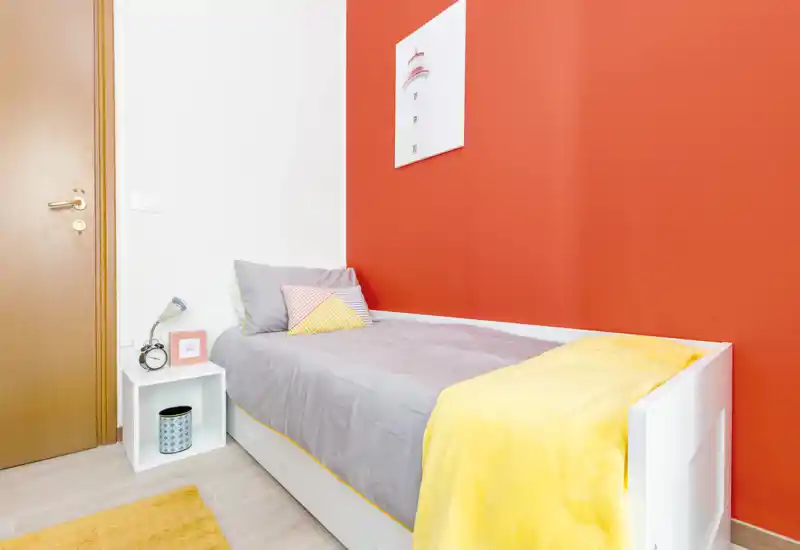 Camera singola luminosa e ordinata con struttura letto bianca, biancheria grigia e un caloroso plaid giallo su una parete accentata color corallo — ideale per un soggiorno accogliente.
