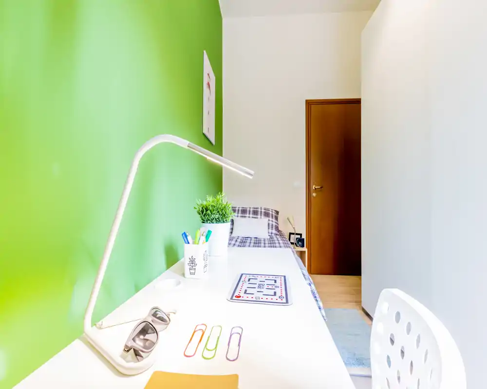 Camera singola luminosa e ordinata con scrivania bianca, parete verde e letto ben rifatto — ideale per studenti o una persona sola.