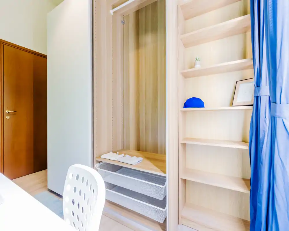 Angolo della camera luminoso con un armadio aperto in legno dotato di ripiani e cassetti a rete, una sedia da scrivania sul bordo e una tenda blu; buona luce naturale e stile pulito e minimale.