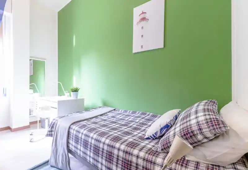 Camera da letto singola luminosa con copriletto a quadri, cuscini decorativi e scrivania vicino a una grande finestra; parete verde a contrasto e arredo minimal creano un ambiente tranquillo e accogliente.